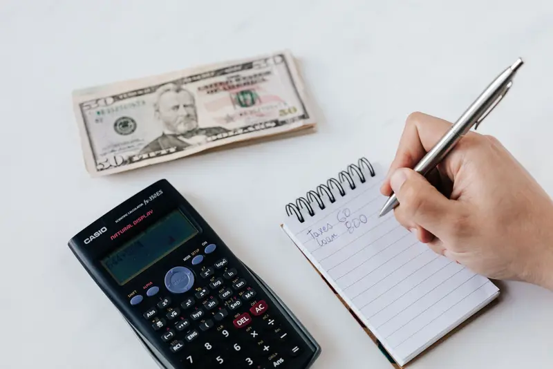 Hand writing figures in a notebook next to a calculator and cash